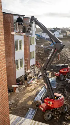 Mand står hævet på kran foran et vindue på øverste sal i et alment lejlighedskompleks med solen i baggrunden